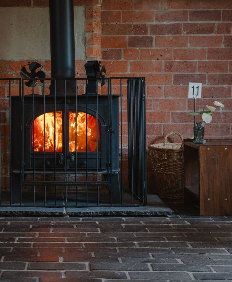 A cast iron wood stove with a glass door and a lit fire, enclosed by a black metal safety guard in front of a brick wall.